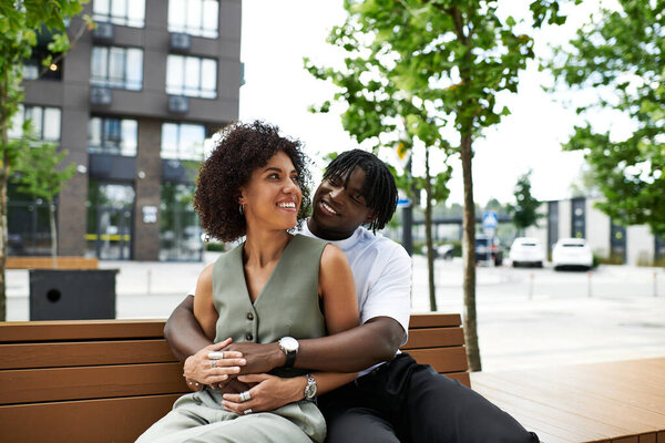 Радостная молодая пара обнимается на скамейке, разделяя улыбки в оживленном городе в течение лета.