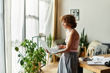 Genç bir kadın dizüstü bilgisayarıyla duruyor, yaz boyunca şık dairesinde işine odaklanmış..