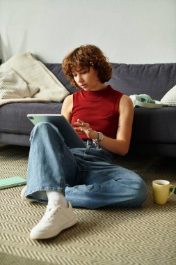 Kıvırcık saçlı genç bir kadın tabletine dalmış, yerde oturuyor..