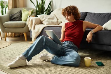 Kıvırcık saçlı genç bir kadın, rahat dairesinde tabletinde kitap okuyor..