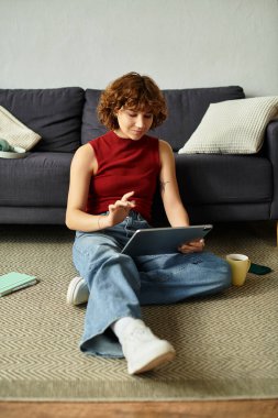 Kıvırcık saçlı kadın kahve içerken tabletini kullanarak rahatça yerde oturuyor..