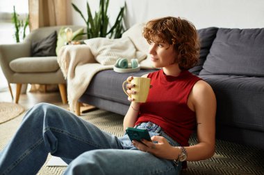 Kıvırcık saçlı kadın modern dairesinde dinleniyor, telefonuna bakarken çayını yudumluyor..