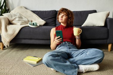 Kıvırcık saçlı kadın yerde oturur, çayını yudumlar ve telefonunu kaydırır, kendini evinde hisseder..