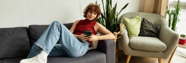 Kıvırcık saçlı kadın rahat bir şekilde uzanıyor. Kapalı alanda yeşillik içindeyken telefonunu kullanıyor.