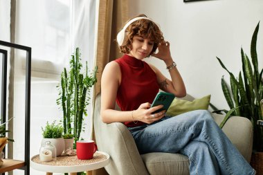 Kıvırcık saçlı genç kadın, telefonuyla müzik dinlerken şık dairesinde dinleniyor..