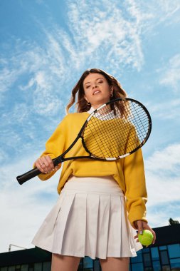 Canlı genç bir kadın tenis için hazır bekliyor, elinde raket ve top, kendinden emin.