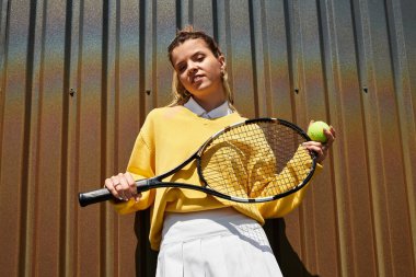 Genç bir kadın tenis raketi ve topuyla rahatça poz verirken yaz atmosferi ışıldıyor..