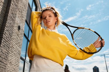 Sarı kazaklı, kendine güvenen genç bir kadın mavi gökyüzünün altında tenis raketiyle poz veriyor..