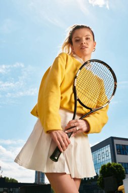 Sarı kazaklı ve beyaz etekli genç bir kadın güneş ışığında tenis raketi tutuyor..