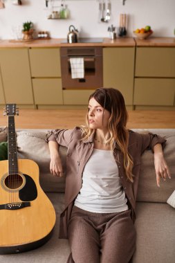 Genç bir kadın sıcak bir mutfakta rahat bir kanepede dinleniyor ve gitar çalıyor..