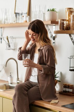 Neşeli genç bir kadın mutfak tezgahında oturup gün ışığında içkisinin tadını çıkarıyor..