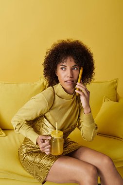 Neşeli sarı bir stüdyoda, kıvırcık saçlı bir kadın içki içerken bir aletin üzerinde konuşur..