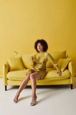 Kıvırcık saçlı bir kadın sarı bir kanepede rahatlar, canlı bir stüdyoda içkisini yudumlar..