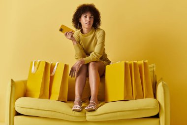 Sarı giyinmiş kıvırcık saçlı bir kadın renkli alışveriş torbalarının arasında dinlenirken akıllı bir telefon tutuyor..