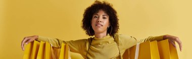 Kıvırcık Afrikalı Amerikalı bir kadın, açık sarı pankarta sarılı alışveriş çantalarını sunar.
