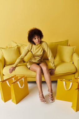 Şık kıvırcık bir kadın, parlak alışveriş torbalarıyla çevrili sarı bir koltukta gelişigüzel oturur..