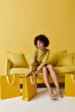 Kıvırcık saçlı siyah bir kadın neşeli bir stüdyo ortamında sarı alışveriş çantalarıyla takılıyor..