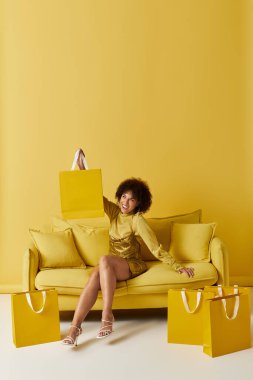 Parlak sarı elbiseli kıvırcık saçlı bir kadın neşeli bir stüdyoda alışveriş torbaları tutuyor..