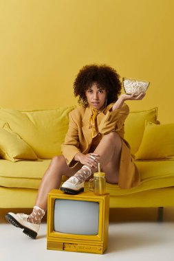 Kıvırcık saçlı şık bir kadın sarı bir kanepede otururken elinde bir kase patlamış mısır tutuyor..