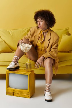 Kıvırcık Afrikalı Amerikalı kadın canlı bir stüdyoda patlamış mısır yerken sarı bir koltukta oturur..