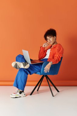 Kıvırcık saçlı, modaya uygun giyinmiş bir kadın laptopuyla parlak bir ortamda etkileşime giriyor..