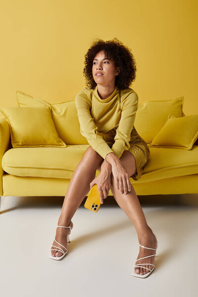 Кудрявая афроамериканка на жёлтом диване вовлекается со своим смартфоном в студию.