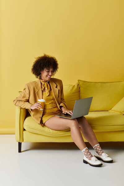 Радостная женщина в стильном желтом ансамбле взаимодействует со своим ноутбуком, наслаждаясь напитком.