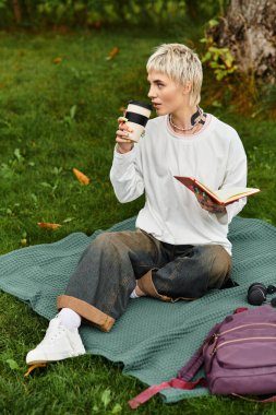 Genç bir kadın battaniyede dinleniyor, kahvesini yudumluyor ve parkta kitap okuyor..