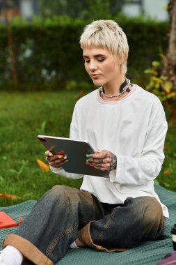 Genç bir kadın açık havayı sever, bir battaniyeye oturur ve tabletine odaklanır..