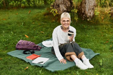 Parktaki bir battaniyeye oturmuş kahvesini yudumluyor ve gülümsüyor..