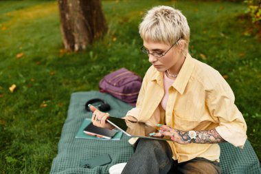 Güzel bir parkta, şık genç bir kadın çimlerin üzerinde otururken tabletiyle meşgul olur.