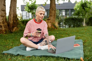 Bir battaniyenin üzerinde rahat bir şekilde oturan şık genç bir kadın kahvesini yudumlarken dizüstü bilgisayarına odaklanıyor..