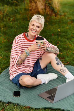 Genç bir kadın kahvesinden zevk alıyor ve yeşil bir parktaki bir battaniyede dizüstü bilgisayarıyla çalışıyor..
