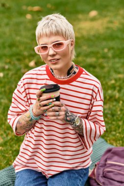 Şık genç bir kadın güneşli bir parkta battaniyede kahve yudumluyor..