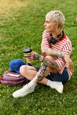 Parkta çimenlerde oturan, kulaklıkları ve dövmeleri olan canlı genç bir kadın..