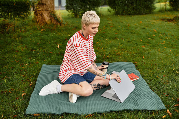 A young woman sits on a green blanket in the park, focused on her laptop while sipping a drink.