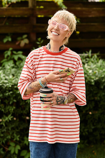 Joyful young woman with tattoos relaxes in a vibrant park, holding a drink and smiling warmly.