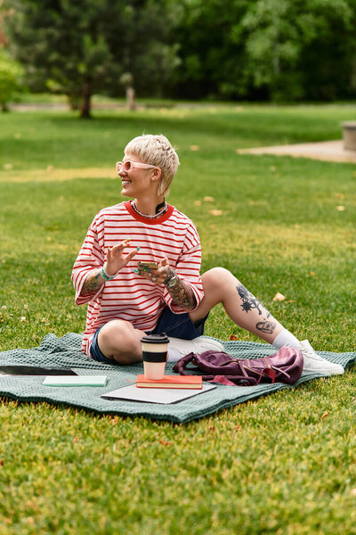 A stylish young woman laughs and sips coffee on a blanket in a lush park.