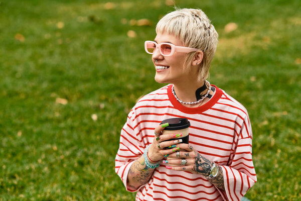 Stylish and cheerful woman sits in a vibrant park holding a cup of coffee while enjoying the day.