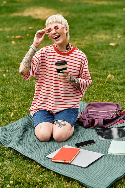 A young, fashionable woman sits on a blanket in a park, laughing joyfully as she enjoys her coffee.
