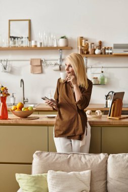 Zarif hatlara sahip son sınıf öğrencisi bir kadın zarif mutfağında elinde telefonla dikiliyor..