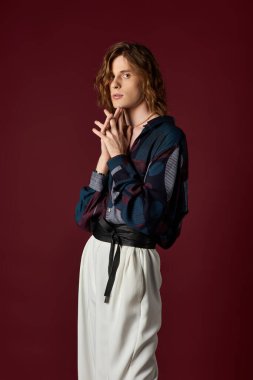 Yakışıklı bir genç adam kendine güvenerek duruyor, sanatsal bir arka planla moda anlayışını ön plana çıkarıyor..