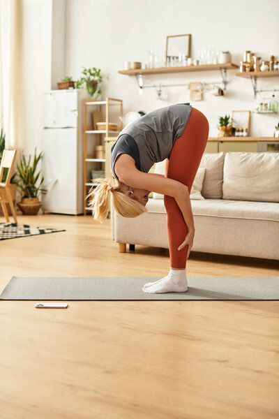 Красивая пожилая женщина изящно наклоняется вперед во время занятия йогой в светлой комнате.