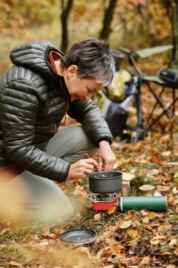 Renkli bir ormanda, yaşlı bir kadın yaprakların arasında taşınabilir bir soba kullanarak yemek hazırlıyor..