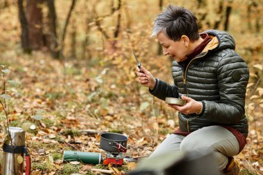 Kıdemli bir kadın orman zemininde diz çöker, sonbahar yaprakları arasında mutlu bir şekilde yemek pişirir..