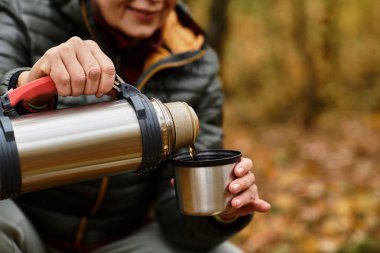 Son sınıf öğrencisi bir kadın renkli sonbahar yaprakları arasında termostan bardağına sıcak bir içecek dolduruyor..