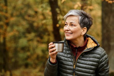 Son sınıf öğrencisi bir kadın, elinde ılık bir içecekle neşeyle gülümsüyor. Etrafı renkli sonbahar yapraklarıyla çevrili..