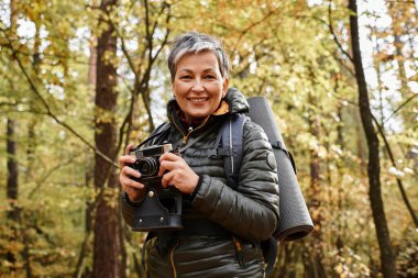 Son sınıf öğrencisi bir kadın, canlı bir sonbahar yeşilliğinin ortasında elinde kamerayla neşeyle gülümsüyor..
