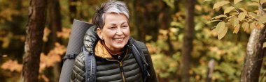 Kıdemli bir kadın, sonbahar renkleriyle çevrili bir orman yolunda yürürken ışıl ışıl gülümser..