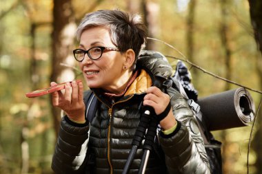 Kıdemli bir kadın, sonbahar yapraklarıyla çevrili güzel bir ormanda yürürken gülümsüyor..